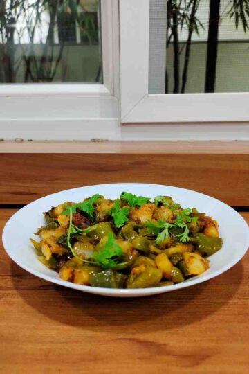Aloo Capsicum Sabzi with chunks of potatoes and bell peppers, garnished with fresh coriander leaves, displayed in a white bowl. The vibrant colors of the vegetables and spices make the dish visually appealing.