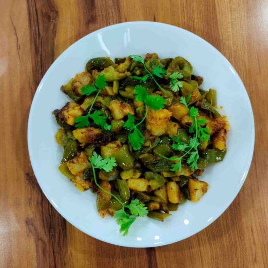 Aloo Capsicum Sabzi with chunks of potatoes and bell peppers, garnished with fresh coriander leaves, displayed in a white bowl. The vibrant colors of the vegetables and spices make the dish visually appealing.